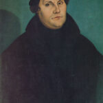 Portrait of a man in a black cloak and hat, hands clasped, against a teal background.