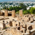 Ancient stone ruin with tall, weathered columns in the foreground and a modern city beyond among trees and hills.