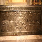 Ornate silver reliquary with carved biblical figures inside a glass display case in a gold-accented church setting