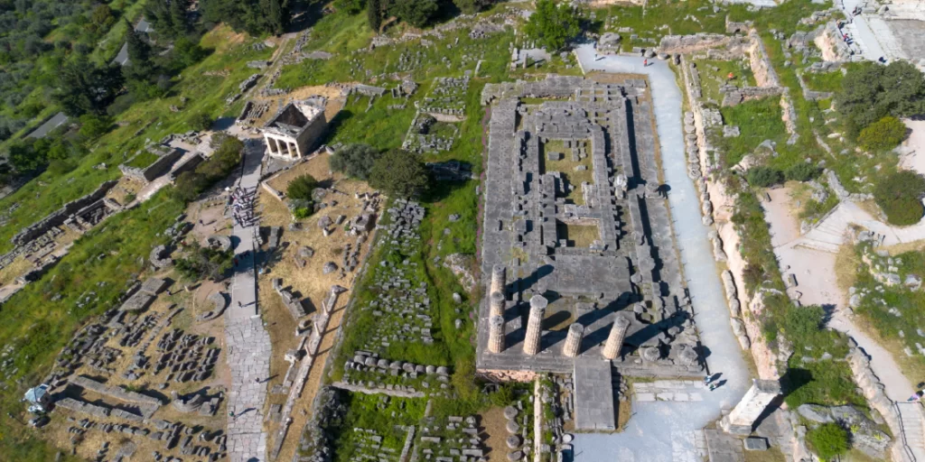 Vista aerea drone del sito archeologico di Delfi in Grecia: rovine del Santuario di Apollo con il Tempio di Apollo al centro, il Tesoro degli Ateniesi in alto a sinistra, la Via Sacra che serpeggia tra i resti delle fondamenta, colonne doriche ancora in piedi e blocchi di pietra sparsi sul pendio del Monte Parnaso