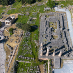Aerial view of ancient ruins with a rectangular stone complex, tall columns, and surrounding grassy, rocky landscape.