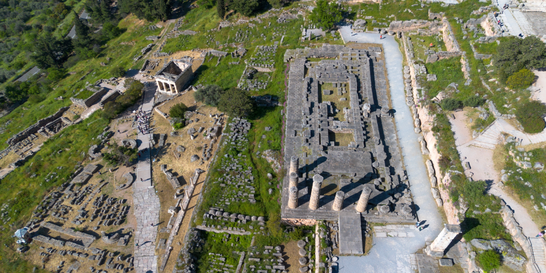 Aerial view of ancient ruins with a rectangular stone complex, tall columns, and surrounding grassy, rocky landscape.