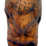 Orange-brown carved mask with closed eyes and a subtle smile, resembling a tribal face.