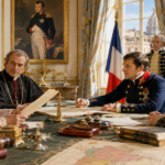 Group of dignitaries in 19th-century attire gathers around a map-covered table, signing documents in a gilded, ornate room.