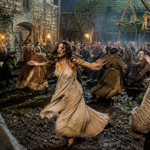 Central woman in a flowing beige dress dances with arms outstretched as villagers whirl around at night in a torchlit medieval square; muddy ground.