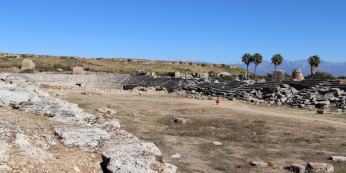 Perge Ancient stone amphitheater with tiered seating under a clear blue sky; a few visitors walk in the arena area