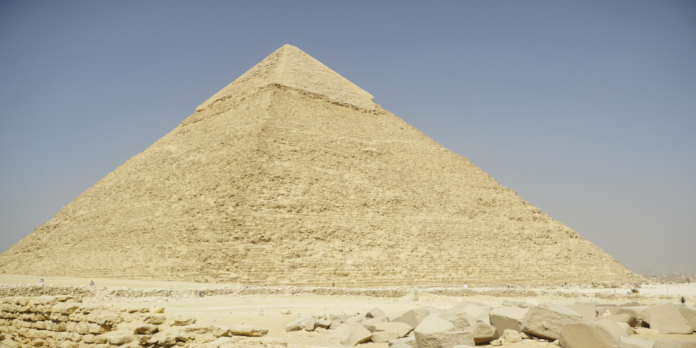 Piramide di Cheope Great Pyramid of Giza under a clear blue sky, with a sandy foreground and scattered stones in view.