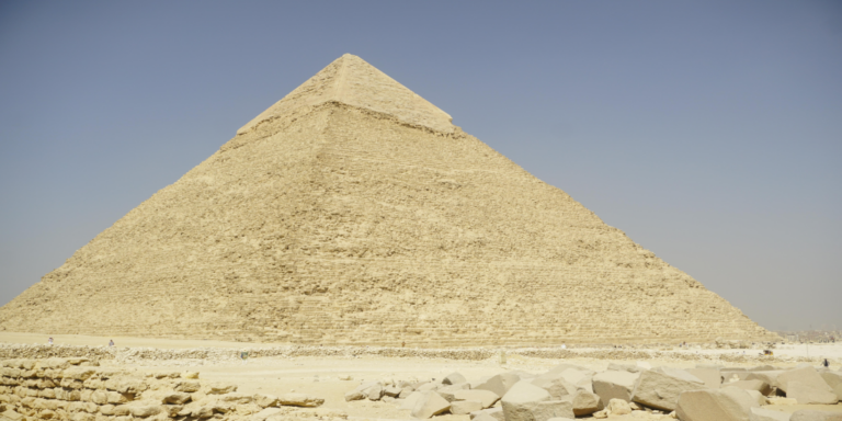 Great Pyramid of Giza under a clear blue sky, with a sandy foreground and scattered stones in view.