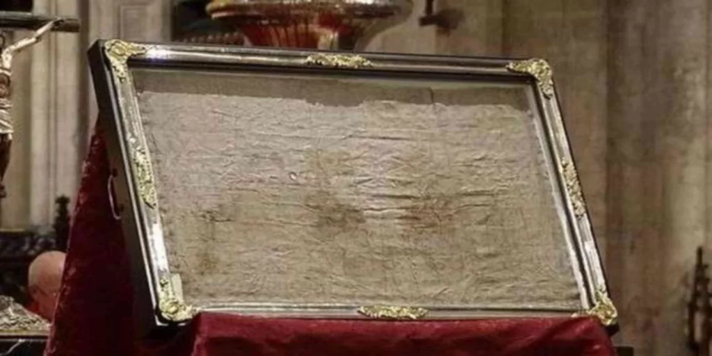 Il Sudario di Oviedo esposto nella Cámara Santa della cattedrale di San Salvador: telo di lino rettangolare color bruno chiaro con macchie ematiche visibili, conservato in una teca con cornice dorata e bordi argentati, poggiata su un cuscino di velluto rosso durante una cerimonia di ostensione