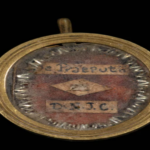 Gold round pendant with a small loop at the top, revealing a worn coin-like inset with illegible handwritten labels inside a glassy cover.