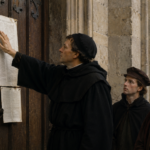 A man in a black monk’s robe points to a large parchment posted on a wooden door, as four others in period clothing look on outside a stone building.