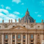 Facade of St. Peter's Basilica with statues along the roofline and a large central dome under a bright blue sky