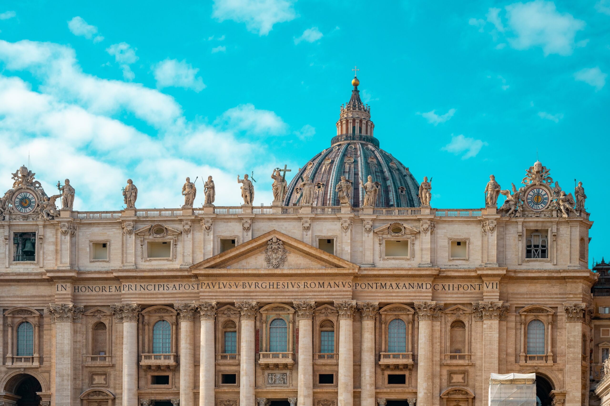 Facade of St. Peter's Basilica with statues along the roofline and a large central dome under a bright blue sky