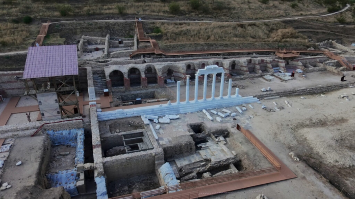 Archaeological site with ancient ruins, stone walls, arches, and a white colonnade in the center.