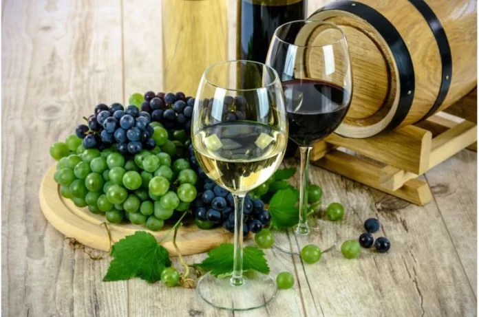 Two wine glasses (white and red) sit on a wooden table beside green and purple grapes and a wine barrel in the background.