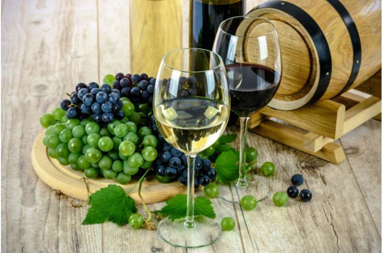 Two wine glasses (white and red) sit on a wooden table beside green and purple grapes and a wine barrel in the background.