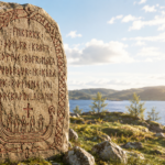 Large standing stone carved with runic inscriptions and a boat-shaped relief at the base, set beside a rocky lakeshore under a blue sky.