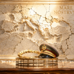 Ceremonial military cap with a gold laurel wreath sits on a metal stand before a cracked marble map relief and the inscription 'MARE NOSTRVM'.