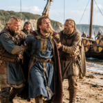 Bearded Viking warriors in blue and brown tunics escort a man in a blue tunic ashore from a longship on a sandy beach, shields and sea behind them.
