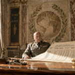 Ufficiale fascista in uniforme militare consulta documenti del Ministero delle Corporazioni alla scrivania, con iscrizioni 'Roma o Morte' e 'Dux e Impero' sullo sfondo, Roma
