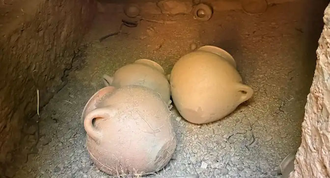News-20260616-Italy-Etruscan-Tomb Three dusty clay pots with small loop handles sit on a cracked dirt floor inside a dark clay kiln or storage chamber by a wall of earth.