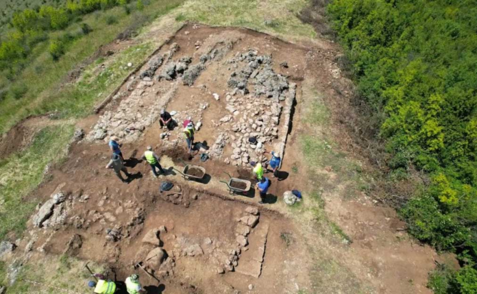 News-20260617-Albania-Temple Aerial view of archaeologists excavating a rectangular site with stone walls and wheelbarrows on a dirt field surrounded by trees.