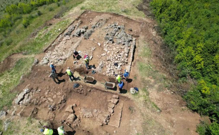 Aerial view of archaeologists excavating a rectangular site with stone walls and wheelbarrows on a dirt field surrounded by trees.