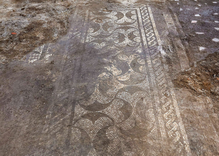 Ancient decorative mosaic fragment with intricate white patterns exposed in dirt at an excavation site.