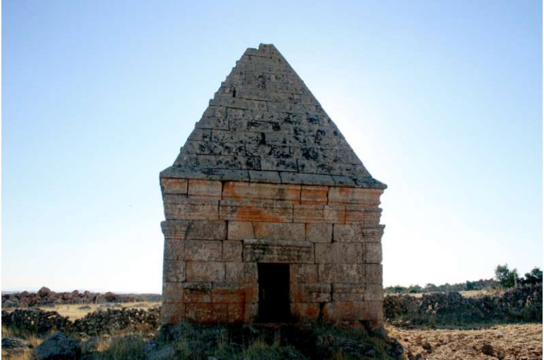 Stone chapel ruin with a pyramidal roof and a dark doorway, set in a dry, open landscape.