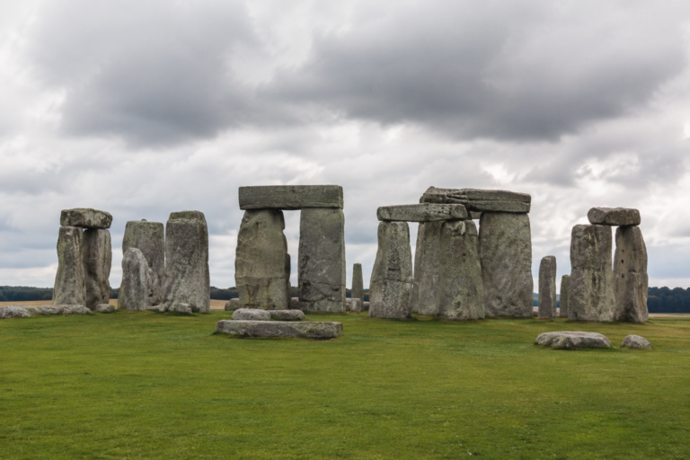 Stonehenge-style monument: a circle of large upright stones with horizontal lintels on a grassy field under a cloudy sky