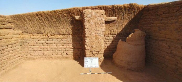 Egitto Archaeological trench with sun-baked mud-brick walls, a central ruined pillar, a whiteboard with notes, and a measurement scale on the sandy floor.