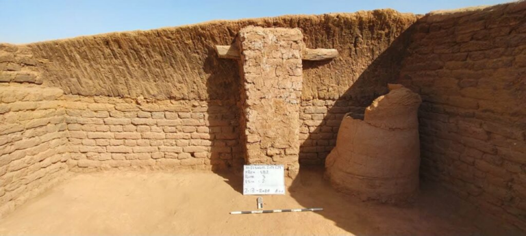 Archaeological trench with sun-baked mud-brick walls, a central ruined pillar, a whiteboard with notes, and a measurement scale on the sandy floor.