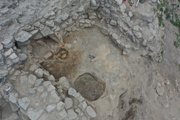Top-down view of an archaeology dig showing circular stone-lined pits in a dirt trench surrounded by rubble walls and soil.