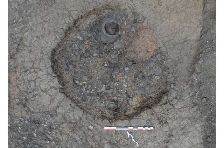 Open excavation exposing a circular metal feature in cracked asphalt, with a measurement scale at the bottom for reference.