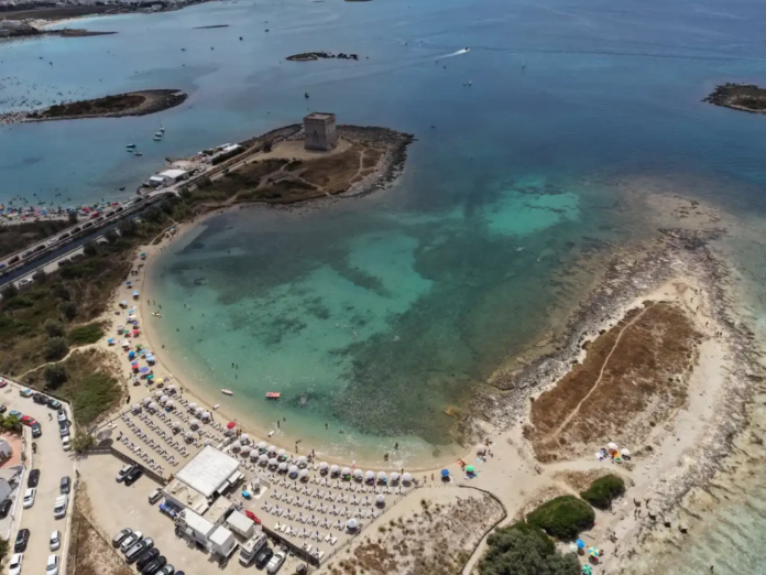 Aerial view of a sandy bay with turquoise water, crowded beach umbrellas, and a small island with a square tower connected by a causeway.