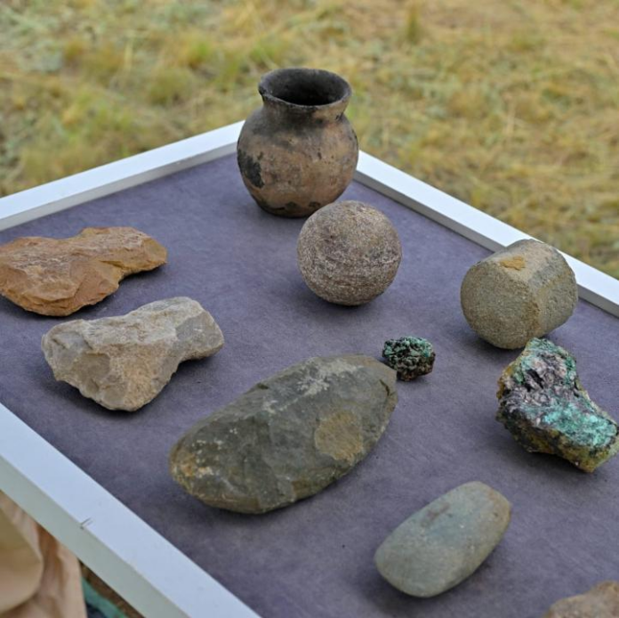 Display of assorted rocks and minerals on a purple-framed tray, including a ceramic pot, round stone sphere, and several rough rocks outdoors