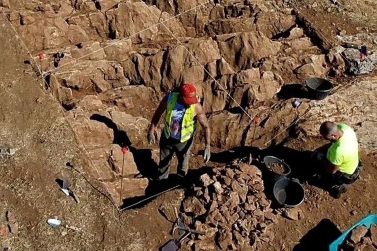 Archaeologists in bright vests working in a rocky trench, sorting stones and placing markers along string lines for excavation progress.