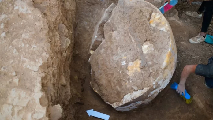 Stele Ninive Large, rounded boulder partially buried in a dirt trench during an excavation, with a person brushing soil nearby wearing gloves and sneakers and a blue arrow marker in the foreground.