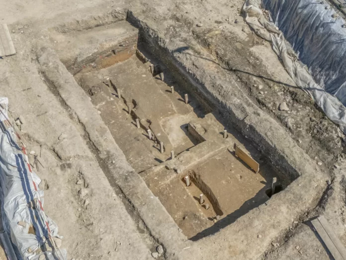 Switzerland-Vindonissa-Roman-Campjpg Archaeological excavation pit with stepped trenches, wooden stakes, and exposed soil at a dig site.