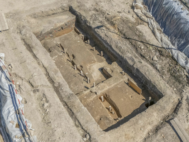 Archaeological excavation pit with stepped trenches, wooden stakes, and exposed soil at a dig site.