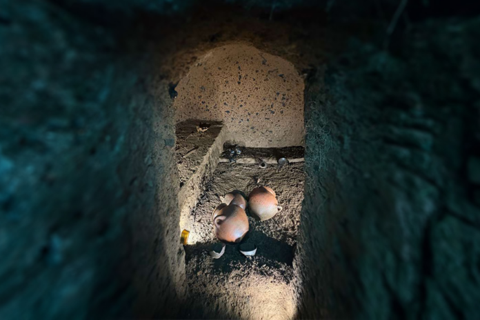 Inside a dark earthen tunnel or shaft, with curved walls, showing a cluster of pale pink, round objects on the dusty floor beneath a beam of light.