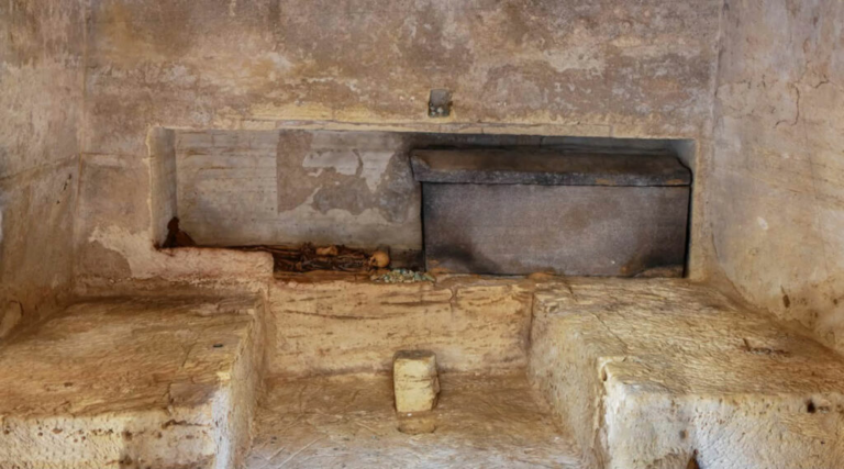 Interior of a rustic stone oven or kiln set into a plastered wall, with a metal door and surrounding stone benches.