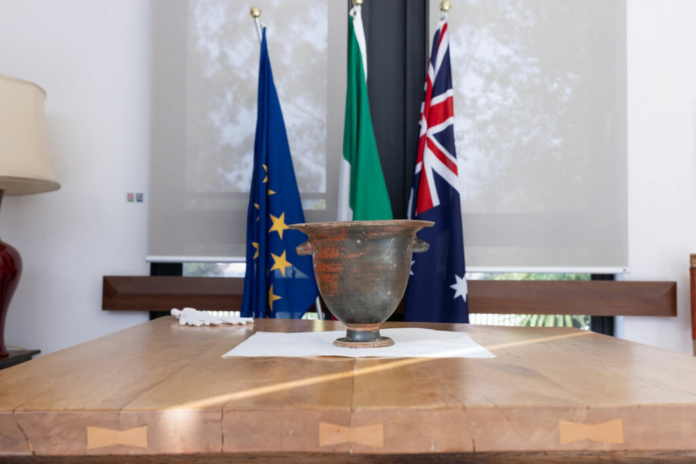 Rusty antique metal chalice centered on a wooden table with EU, Ireland, and Australia flags in the background.