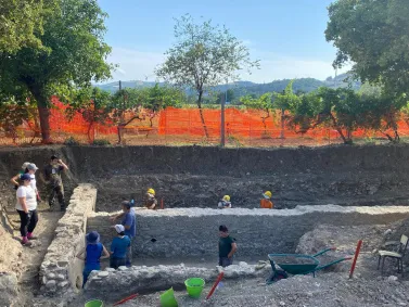 Villa romana Children in yellow hard hats explore a stone-lined excavation pit supervised by adults outdoors.