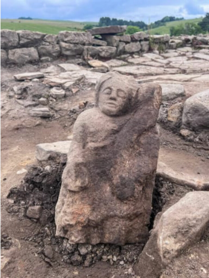 Stone sculpture of a seated, serene figure with eyes closed, resting against its arm at an archaeological site with stone walls behind it in a rural landscape.