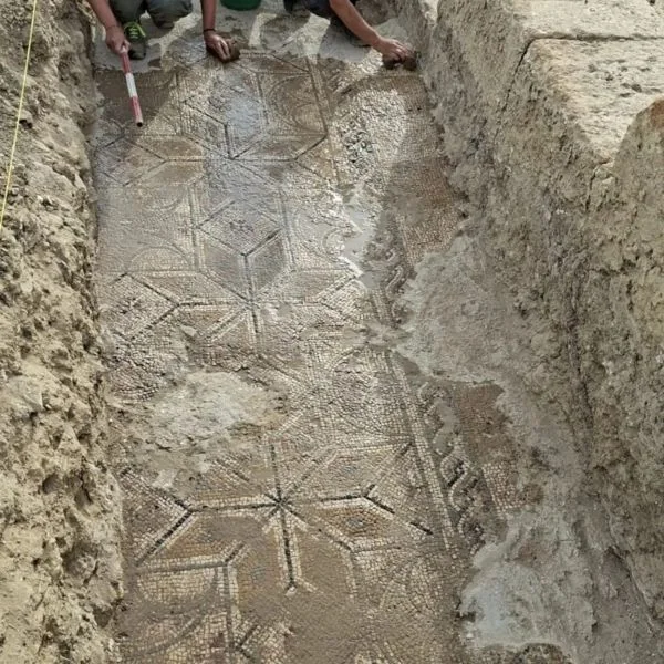 Agrigento Archaeologists uncovering a geometric mosaic floor in a trench, showing star and hexagon patterns in wet soil.