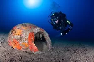 Anfora Scuba diver swimming near an orange rusted underwater pipe with a dark tunnel opening on a sandy seabed.