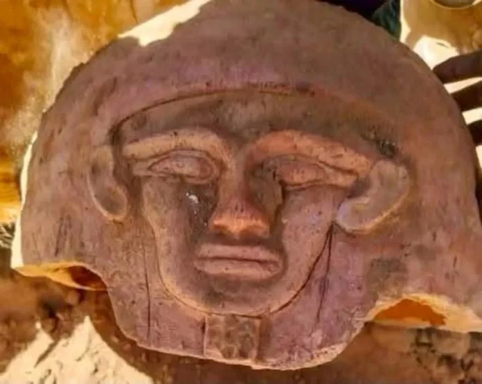 Bahariya Ancient carved stone relief of a human face with large rounded ears and a helmet-like headpiece, weathered brown-red stone with a chipped edge at the bottom.