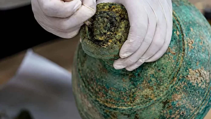 Gloved hands carefully handling a small, heavily patinated green bronze vessel during conservation.
