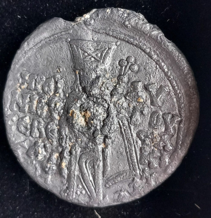 Ancient circular coin with a standing figure in the center, holding a tall staff or scepter, surrounded by worn inscriptions and decorative marks on a rough, pitted surface.